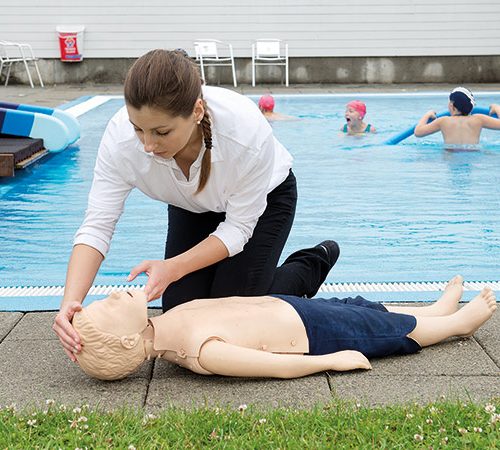 Kvinna sitter vid pool med en HLR-docka i barnstorlek på backen. Barn-HLR