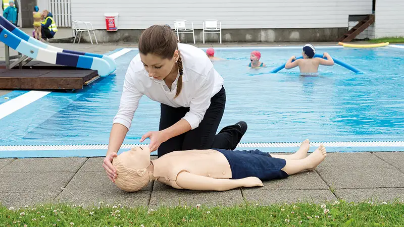 Kvinna sitter vid pool med en HLR-docka i barnstorlek på backen. Barn-HLR
